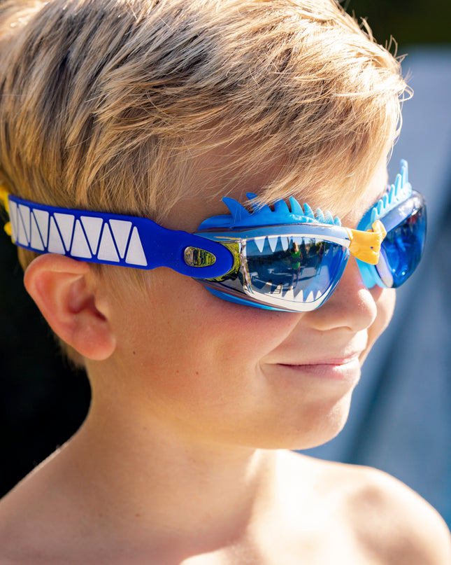 boy smiling wearing draco the dragon kids' swim goggles in blue dragon showing a silicone spiked gasket, sharp teeth printed lens and patterned strap