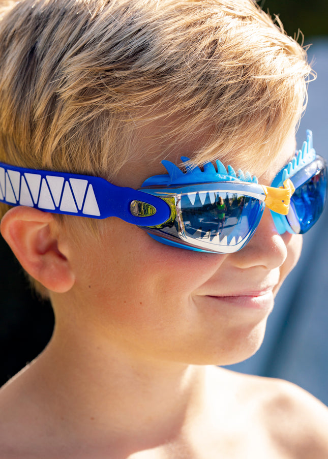 boy smiling wearing draco the dragon kids' swim goggles in blue dragon showing a silicone spiked gasket, sharp teeth printed lens and patterned strap