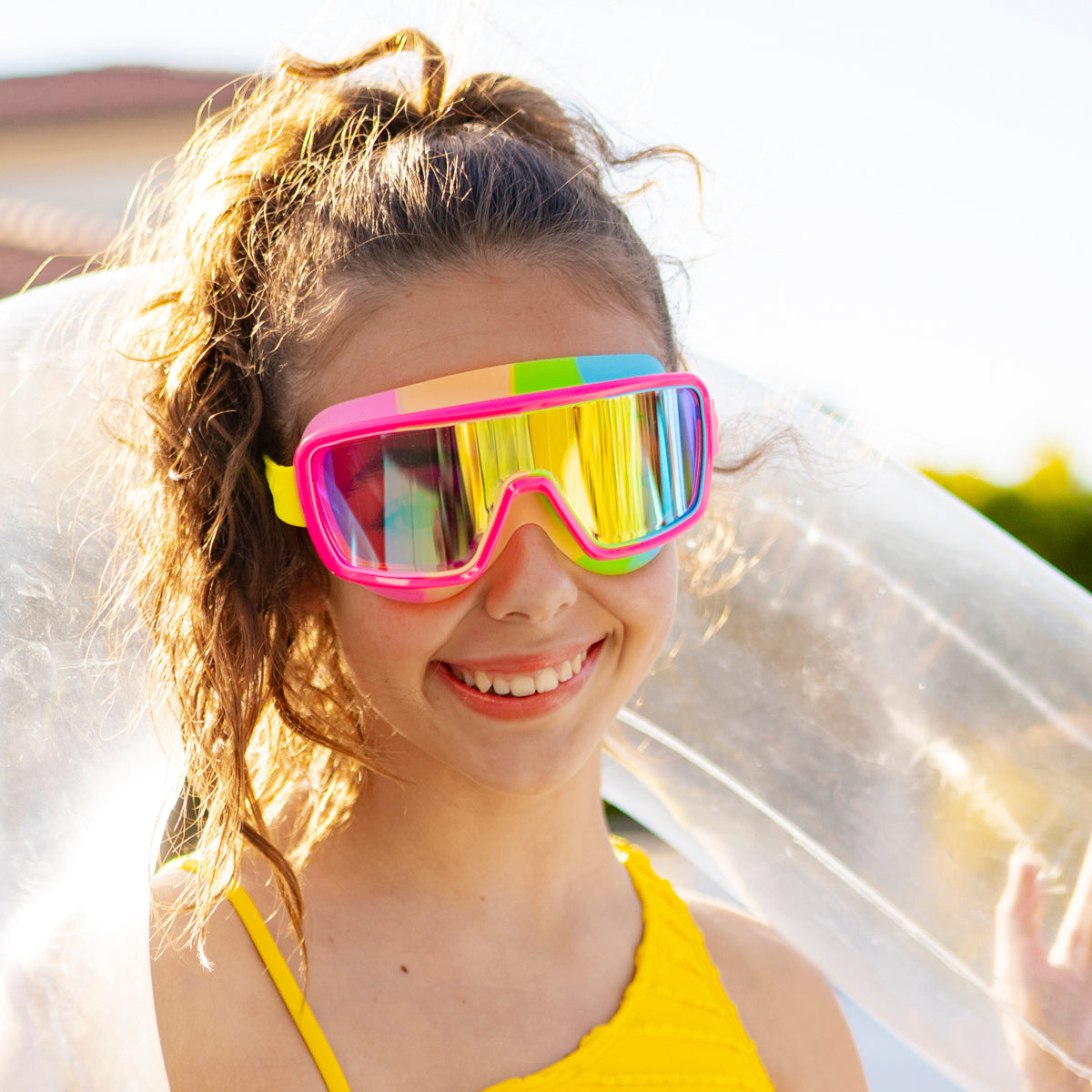 girl smiling poolside wearing chromatic swim goggles in spectro strawberry showing a hot pink frame and rainbow colored gasket with reflective lens