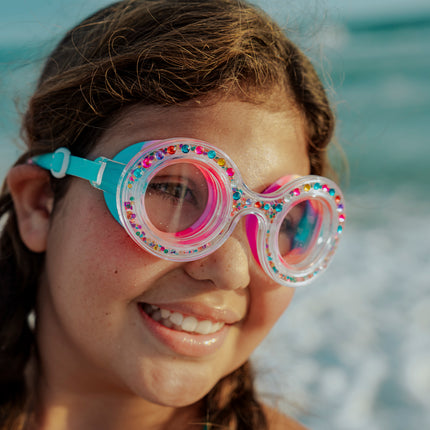 girl at beach in front of ocean smiling wearing dazzling kids' round shaped swim goggles in gumball gleam showing rhinestones and 3D rhinestones in the double walled frame
