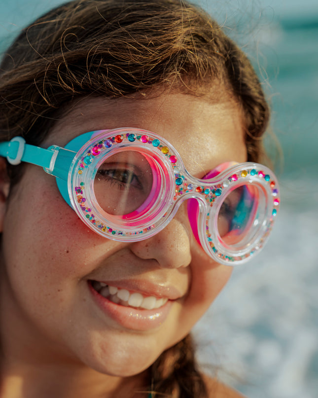 girl at beach in front of ocean smiling wearing dazzling kids' round shaped swim goggles in gumball gleam showing rhinestones and 3D rhinestones in the double walled frame