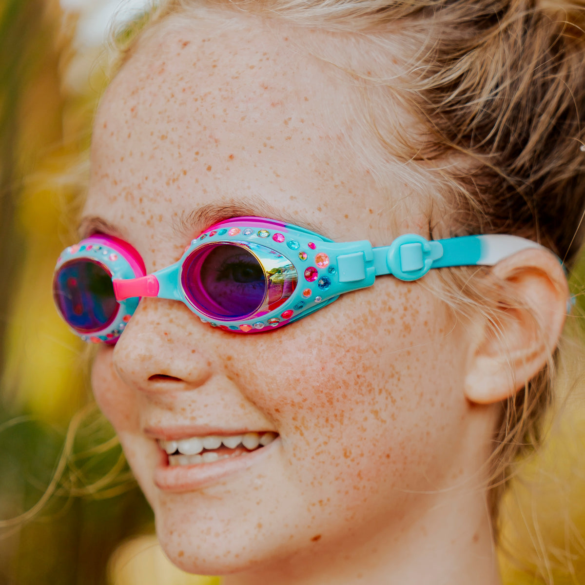 close-up view of girl smiling wearing glimmering gemstones kids' swim goggles in crystal violet showing colorful rhinestones on a bright blue frame