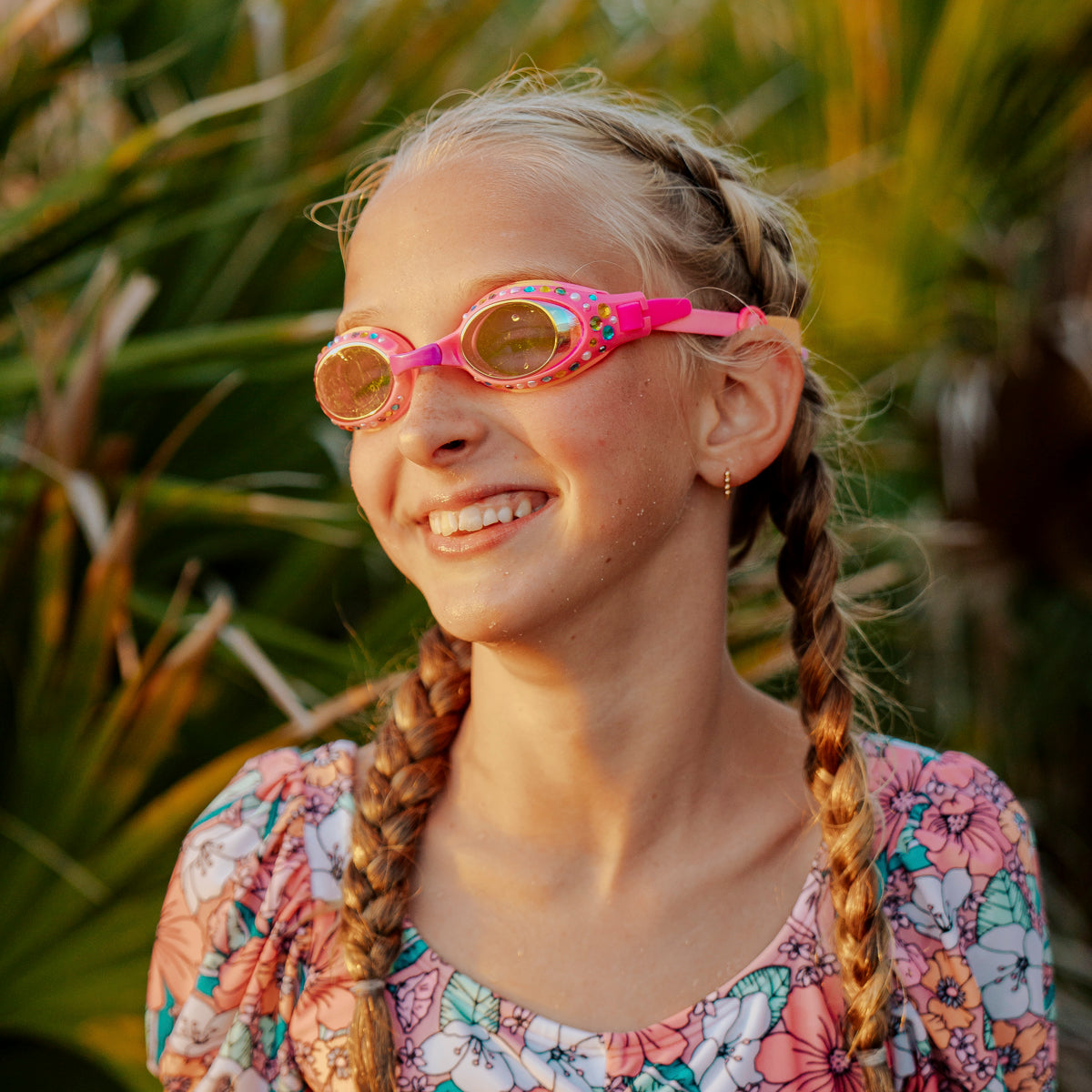girl smiling poolside wearing glimmering gemstones kids' swim goggles in tropical tanzanite showing rhinestones and gems on a bright pink swim goggle frame