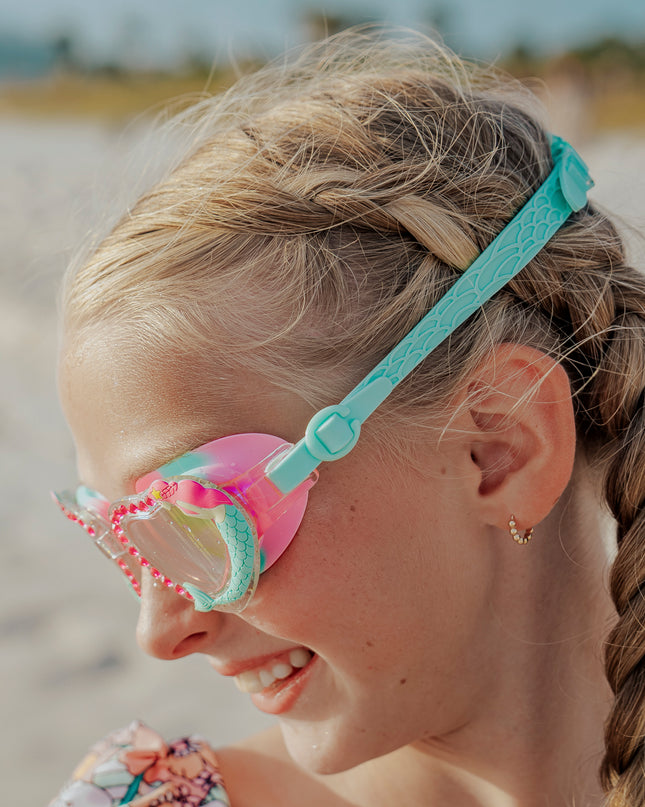 girl at beach looking down wearing mystical mermaid kids' swim goggles showing 3d mermaid detail and rhinestones and fishtail textured strap