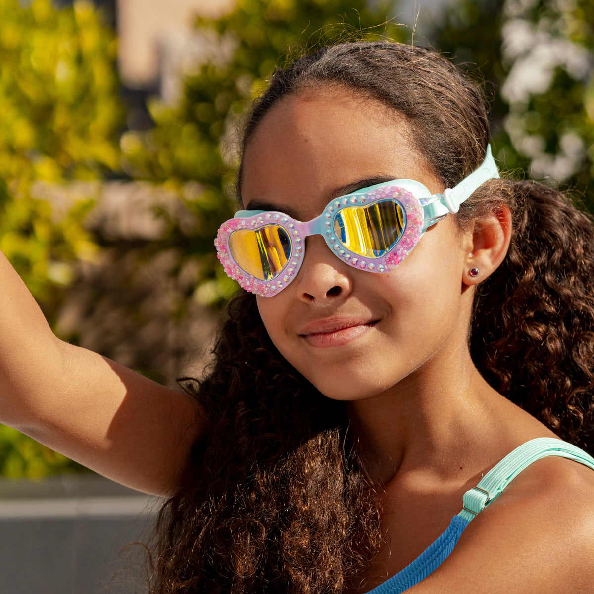 girl smiling outside wearing seaquin kids' heart shaped swim goggles in bluetiful showing details of crushed pink rhinestones and a set of rhinestones 