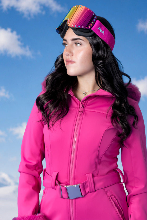 woman standing with blue sky background in pink ski suit wearing powder pop ski goggles in alpen glow
