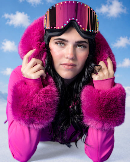 woman laying on snowy ground in pink ski suit wearing powder pop ski goggles in alpen glow