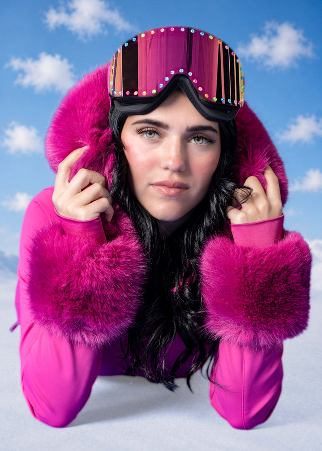 woman laying on snowy ground in pink ski suit wearing powder pop ski goggles in alpen glow