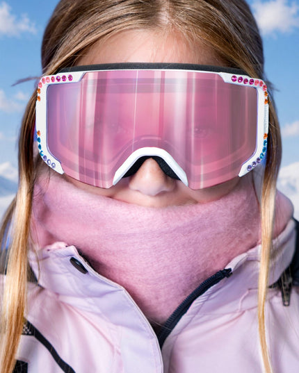 headshot of model wearing pink scarf and jacket matching pink powder pop kids' ski goggles in prism powder showing pink mirrored lens and rainbow rhinestones around white frame