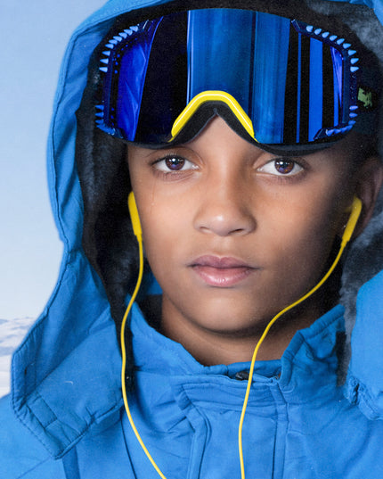 head shot of boy wearing all blue ski jacket and prism peak kids ski goggles in blue bird which features a blue mirrored lens with blue silicone spikes and a yellow nose piece