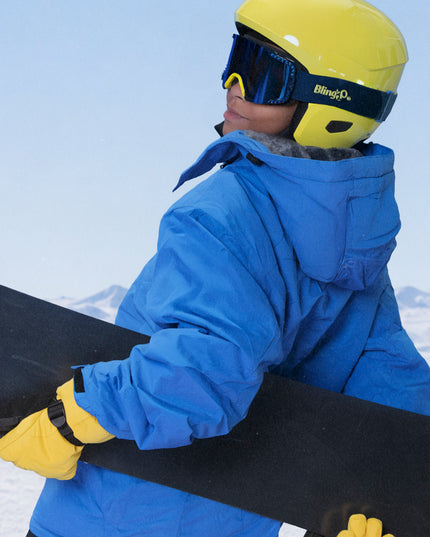 full body shot of boy wearing all blue ski jacket with snowboard and prism peak kids ski goggles in blue bird which features a blue mirrored lens with blue silicone spikes and a yellow nose piece