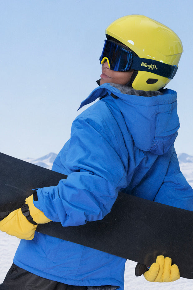 full body shot of boy wearing all blue ski jacket with snowboard and prism peak kids ski goggles in blue bird which features a blue mirrored lens with blue silicone spikes and a yellow nose piece
