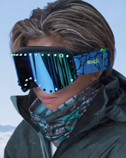 head shot of boy wearing prism peak kids' ski goggles in glacier teal featuring a teal mirrored lens, blue strap, green, white and black silicone spikes with decorated monster eye on either side