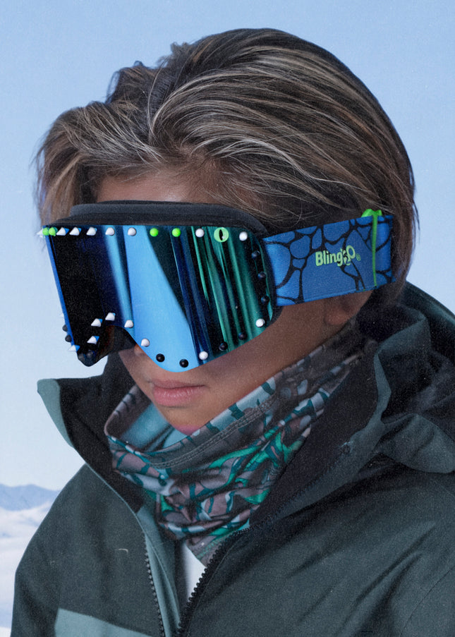 head shot of boy wearing prism peak kids' ski goggles in glacier teal featuring a teal mirrored lens, blue strap, green, white and black silicone spikes with decorated monster eye on either side