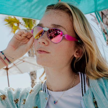 girl posing under sun umbrella wearing hampton beach sunglasses in bright bubblegum showing an aviator frame with mirrored lens and a crown nose piece and mermaid tail themed arms of sunglasses