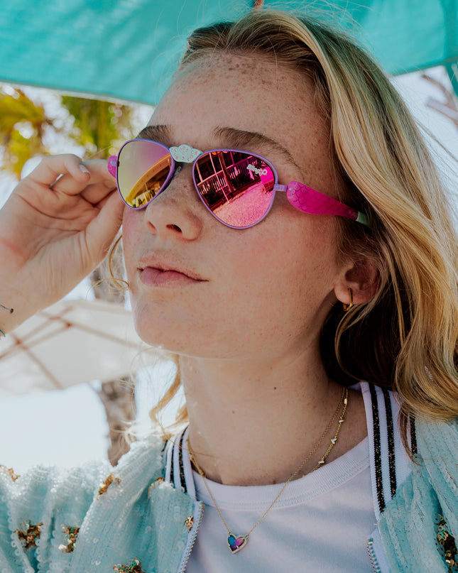 girl posing under sun umbrella wearing hampton beach sunglasses in bright bubblegum showing an aviator frame with mirrored lens and a crown nose piece and mermaid tail themed arms of sunglasses