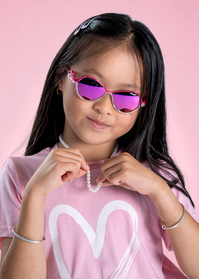 girl posing in pink studio wearing heart beach kids' sunglasses in color rosa romance featuring oval lens with a pink and white glittering frame and reflective lens