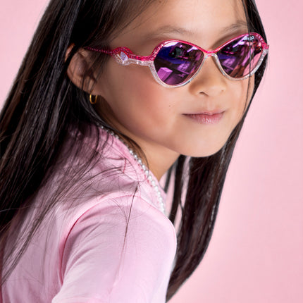 girl posing in pink studio wearing heart beach kids' sunglasses in color rosa romance featuring oval lens with a pink and white glittering frame and reflective lens