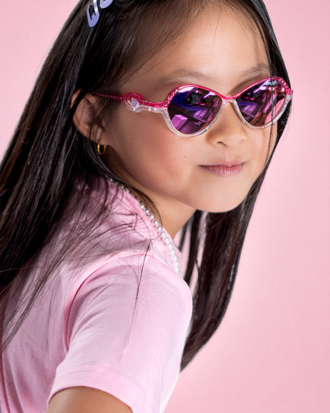 girl posing in pink studio wearing heart beach kids' sunglasses in color rosa romance featuring oval lens with a pink and white glittering frame and reflective lens