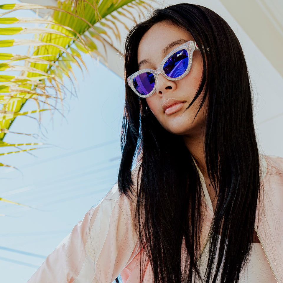 girl posing in sunlight and palm trees wearing malibu beach kids' sunglasses in starship sparkle showing a silver glitter infused frame and pink rhinestones