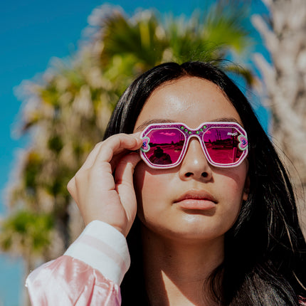 girl posing in sunny weather in front of palm trees wearing miami beach kids' sunglasses in ultra violet showing a purple glitter infused square frame with hearts and rhinestone details