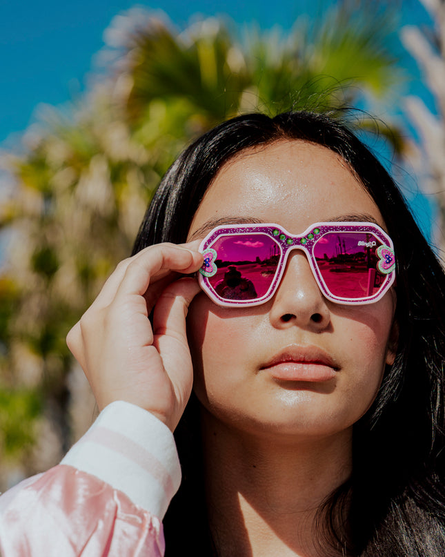 girl posing in sunny weather in front of palm trees wearing miami beach kids' sunglasses in ultra violet showing a purple glitter infused square frame with hearts and rhinestone details