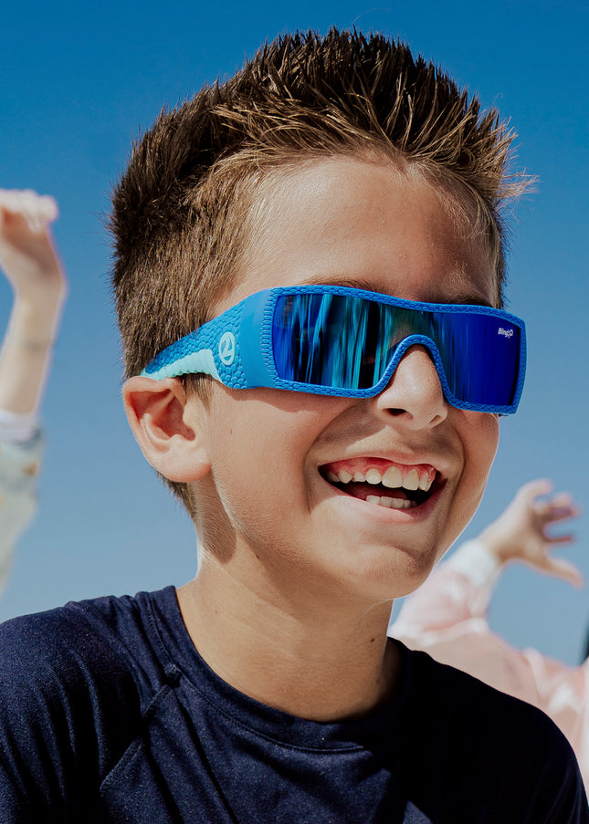 young boy smiling wearing sand harbor kids' sunglasses in sky blue stegosaur showing a sleek frame and dino textured arm of sunglass