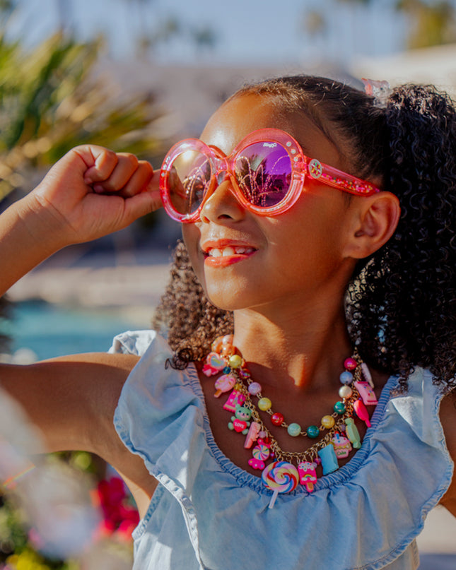 Lifestyle of girl smiling posing wearing glass beach kids sunglasses in blush pink color with 3D gumballs in the arms