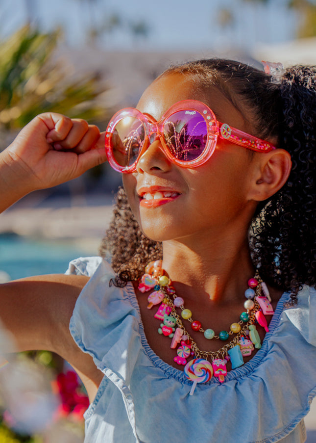 Lifestyle of girl smiling posing wearing glass beach kids sunglasses in blush pink color with 3D gumballs in the arms
