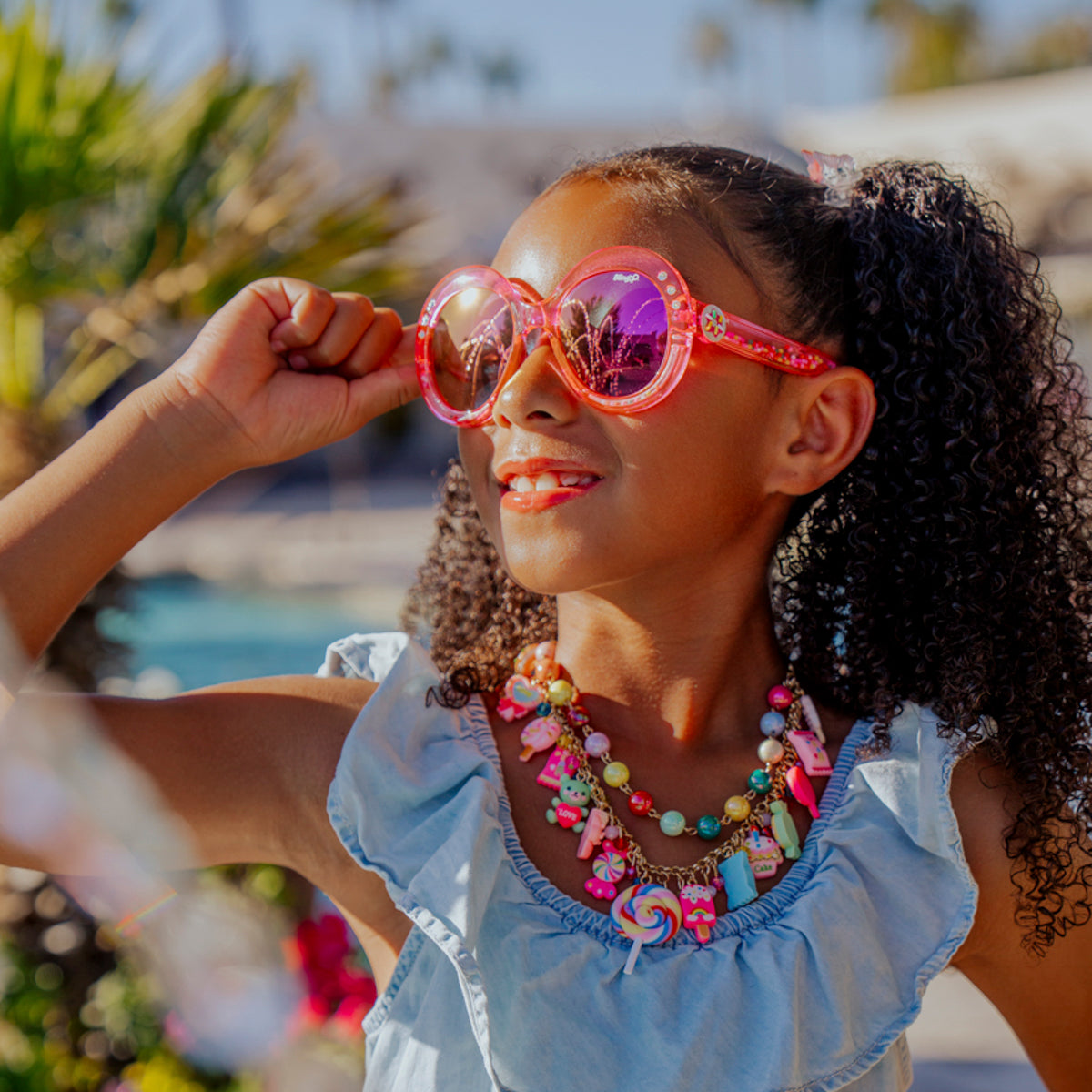 Lifestyle of girl smiling posing wearing glass beach kids sunglasses in blush pink color with 3D gumballs in the arms