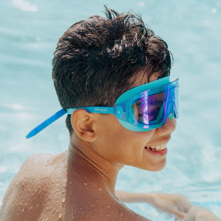 lifestyle of boy smiling sitting in pool wearing hologram kids swim goggles in polar stream blue showing frameless lens and blue colored silicone