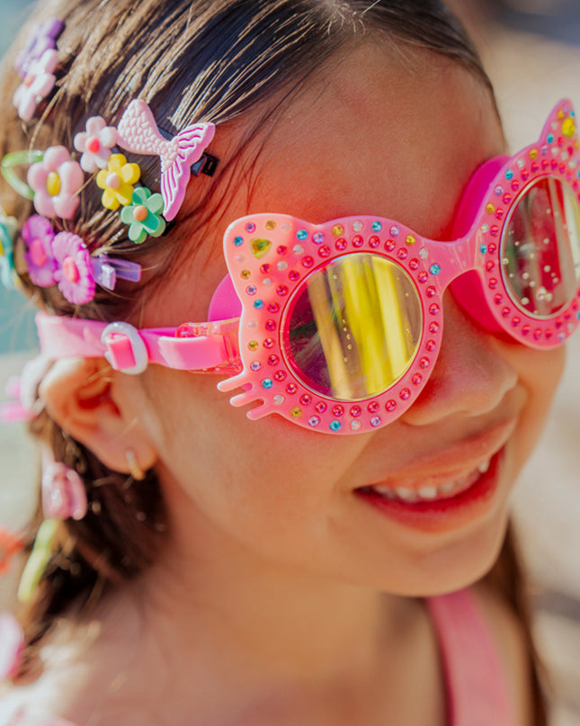 lifestyle of girl smiling in bedazzled accessories wearing Kitten kids swim goggles in mango mittens hot pink cat shaped swim goggles 