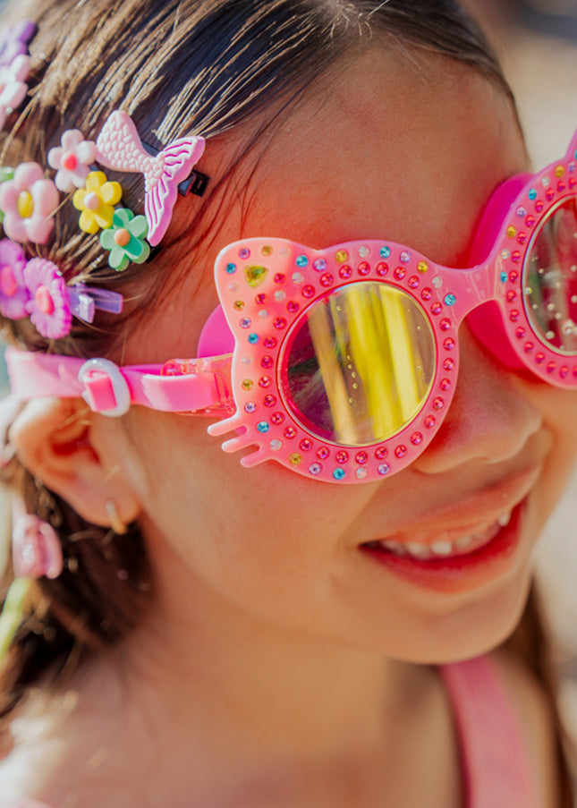lifestyle of girl smiling in bedazzled accessories wearing Kitten kids swim goggles in mango mittens hot pink cat shaped swim goggles 