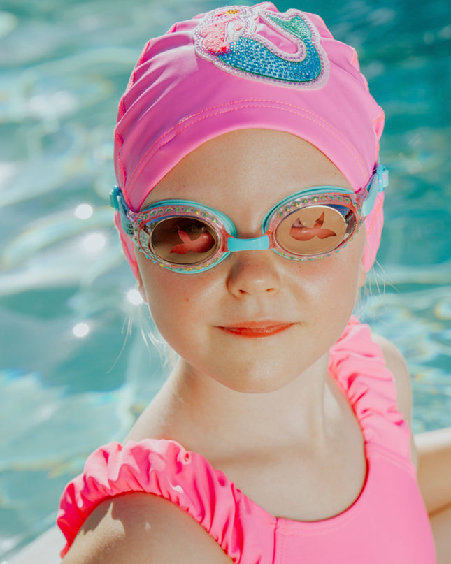 girl in pool wearing mermaid classic kids swim goggles in blue sushi showing rhinestones and translucent mermaid tail printed on lens also wearing mermaid swim cap showing a rhinestone mermaid patch