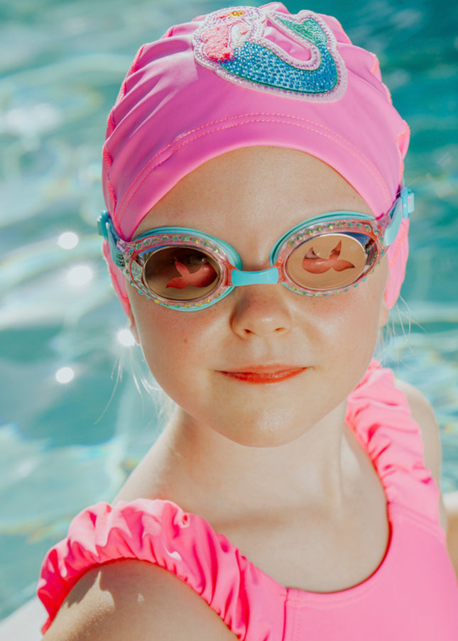 girl in pool wearing mermaid classic kids swim goggles in blue sushi showing rhinestones and translucent mermaid tail printed on lens also wearing mermaid swim cap showing a rhinestone mermaid patch