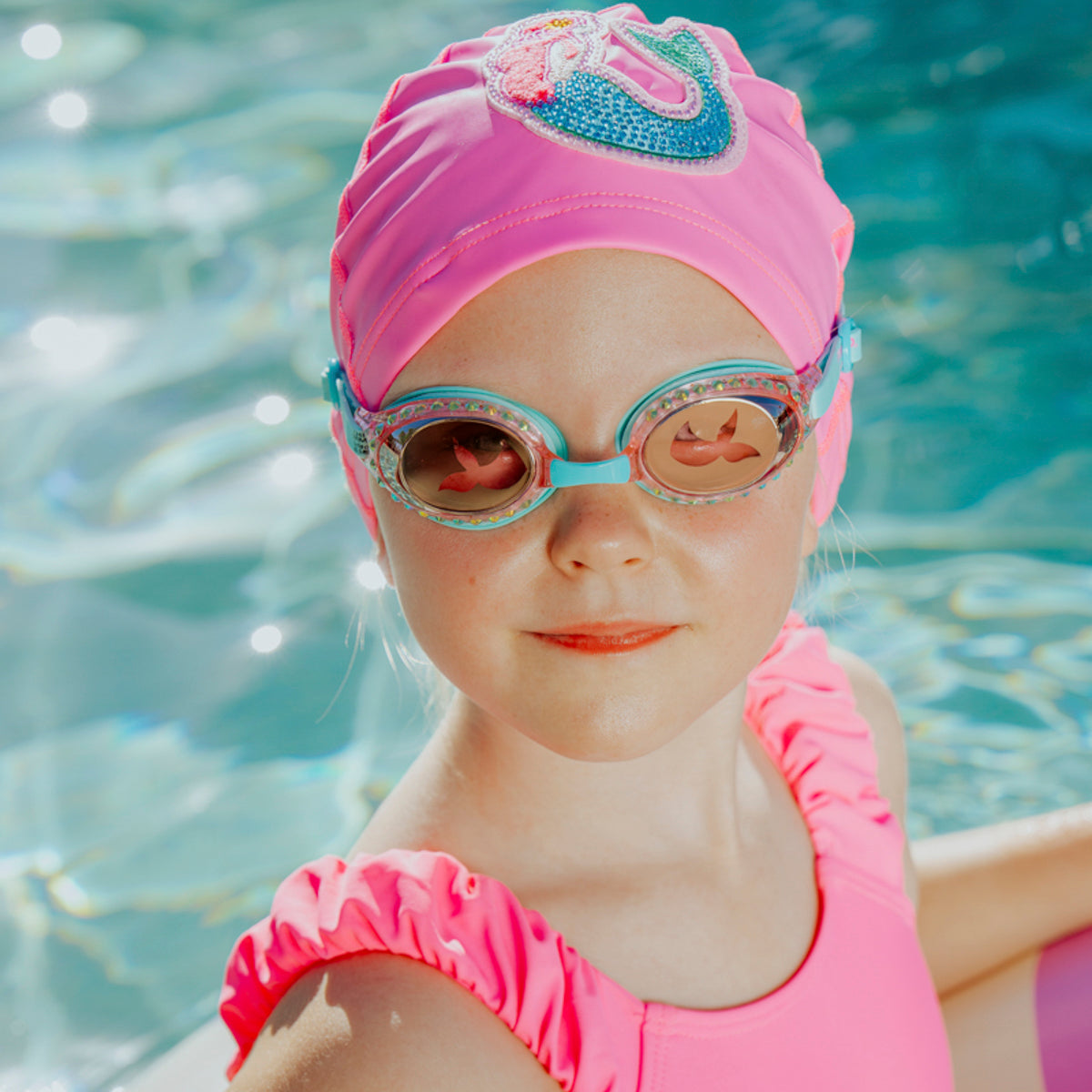 girl in pool wearing mermaid classic kids swim goggles in blue sushi showing rhinestones and translucent mermaid tail printed on lens also wearing mermaid swim cap showing a rhinestone mermaid patch