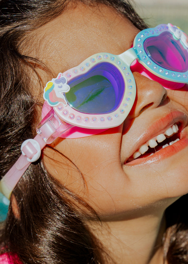 girl smiling and laughing wearing uniglow kids heart shaped swim goggles showing a pastel rainbow coloring, rhinestones and unicorn detail