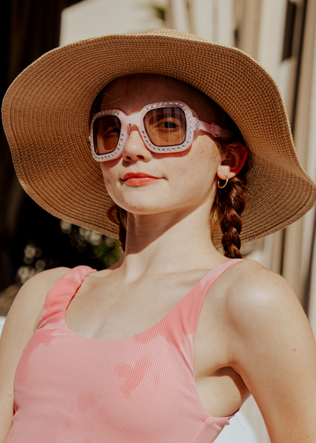 teen girl sitting poolside wearing vibrancy swim goggles in rose quartz showing a pale pink large square frame, rhinestones and tined lens